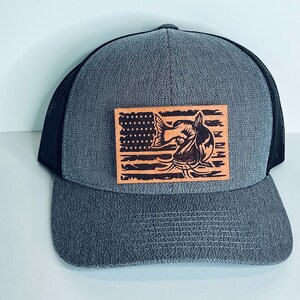 May include: A gray and black trucker hat with a brown leather patch featuring a distressed American flag with a catfish jumping out of the water.