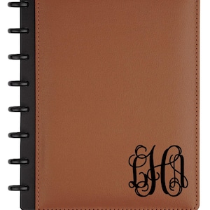 May include: A brown leather-bound notebook with a black spiral binding. The cover features a black monogram in a decorative font. The notebook is rectangular and has a smooth texture.