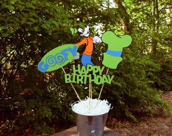 Könnte beinhalten: Ein grünes und blaues Goofy-Geburtstags-Mittelstück mit einem Goofy-Cartoon, der ein Surfbrett mit der Aufschrift "Goofy" und einen grünen Hut hält. Das Mittelstück enthält auch ein Banner mit der Aufschrift "Happy Birthday".