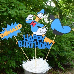 May include: A set of three blue and yellow birthday party decorations featuring Donald Duck, a blue hat, and the words "Happy Birthday" and "Donald".