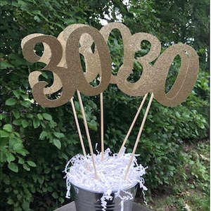 May include: Gold glitter number 30 table centerpiece with four sticks in a silver bucket filled with white shredded paper.