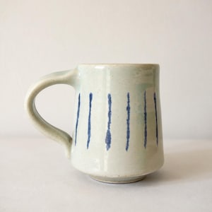 May include: A ceramic mug with a light off-white glaze and a speckled texture. The exterior features a series of vertical, hand-painted dark blue stripes that vary slightly in thickness and intensity, wrapping around the cylindrical body.