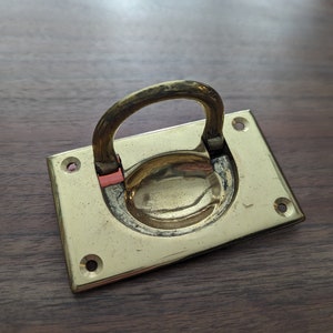 May include: A brass door pull with a rectangular base and a rounded loop handle. The pull has a polished finish and is mounted on a wooden surface.