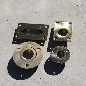 May include: Four brass cabinet pulls in different sizes and shapes. Two are round with a small lip around the edge, one is rectangular with a rounded edge, and one is rectangular with a straight edge.