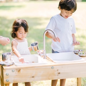 May include: A wooden outdoor play sink with two basins and a chrome faucet. Two children are playing with the sink, using metal cups and small dishes. The scene is set outdoors on a sunny day.