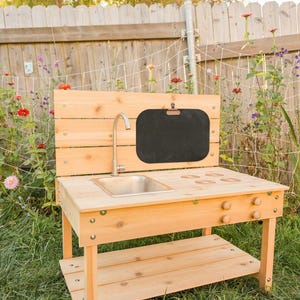 May include: A wooden outdoor play kitchen with a stainless steel sink, a chalkboard, and wooden knobs. The play kitchen is made of light-colored wood and has a lower shelf for storage. It is set up in a grassy backyard with a wooden fence and flowers.