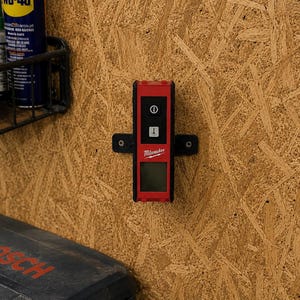 Op de afbeelding: Een rood en zwart Milwaukee laserwaterpas gemonteerd op een muur met een zwarte beugel. Het apparaat is voorzien van een aan-uitknop, een modusknop met een laserpictogram en een digitaal display. De merknaam Milwaukee is in het wit afgedrukt.