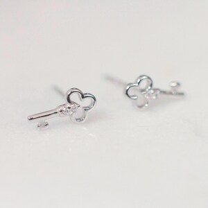 May include: Two silver key-shaped stud earrings. Each earring has a key design with a clover-shaped top and a small decorative element on the key's shaft. The earrings are set against a white background.