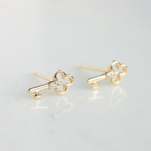 Peut inclure: Une paire de boucles d'oreilles clous en forme de clé de couleur or. Chaque boucle d'oreille présente un motif de clé avec un petit accent de pierres précieuses transparentes. Les boucles d'oreilles sont présentées sur une surface blanche.