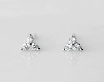 May include: A pair of small, silver stud earrings. Each earring has three clear, round gemstones in a triangular cluster. The earrings are displayed on a white surface with a subtle reflection.