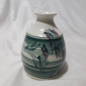 May include: A small white ceramic vase with a green and purple floral design. The vase has a rounded body and a narrow neck.