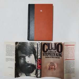 May include: A paperback copy of the Stephen King novel "Cujo" with a red, white, and black cover. The cover features a close-up of a dog's face with sharp teeth. The back cover has a description of the book and a photo of Stephen King.