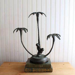 May include: A dark bronze-toned metal candle holder featuring three palm tree structures rising from a circular base. A small monkey figure is sculpted climbing the central palm tree trunk. The base is textured to resemble ground terrain.