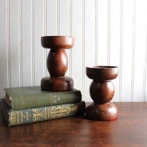 May include: Two dark brown wooden candle holders with a smooth, rounded, stacked design. One candle holder rests on top of two stacked hardcover books, while the other stands directly beside them on a flat surface.
