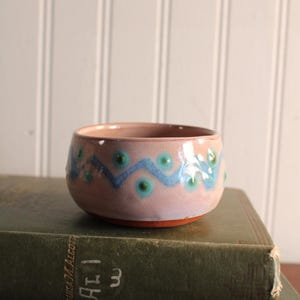 May include: A small, round ceramic bowl with a glossy light pink glaze. The exterior features a hand-painted horizontal blue wavy line pattern accented with small, evenly spaced teal dots. The base of the bowl shows the natural reddish-brown clay.