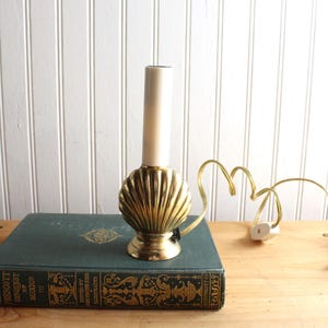 May include: A brass lamp base shaped like a seashell with a vertical white candle-style socket. The lamp features a gold-toned metallic finish with ribbed detailing and includes a thin gold electrical cord with a small white plastic switch.