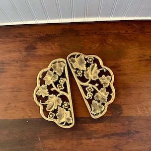 May include: Two ornate, gold-colored metal cabinet pulls with a leaf and flower design. The pulls are in a semi-circular shape and are placed on a dark wooden surface. The background includes a white-painted wood panel.