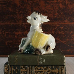 May include: A ceramic donkey figurine with a yellow basket, resting on two vintage green books. The donkey is white with brown and black accents. The books have gold lettering.