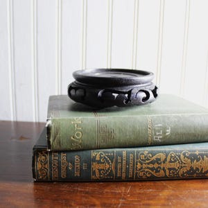 May include: A black circular wooden display stand with carved openwork patterns along the sides, resting on top of two stacked hardcover books titled Work and Conquest of Mexico, featuring green covers with gold lettering and decorative details.