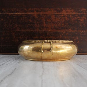May include: A vintage, oval-shaped brass bowl with a hammered texture. The bowl has a decorative rope-like trim around the rim and two small chain accents. The background features a dark brown wooden surface.
