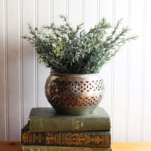 May include: A silver metal planter with a perforated design, filled with green and white faux greenery. The planter sits atop a stack of vintage green books. The background is a white vertical shiplap wall.