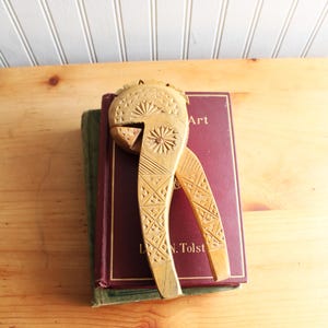 May include: A vintage wooden nutcracker with intricate carved designs, resting on a maroon book with the word "Art" visible. The nutcracker is light brown and features geometric patterns and a floral design.