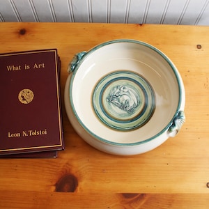 May include: A ceramic bowl with a cream-colored interior and a teal and blue rim. The bowl features a central design with a teal and blue illustration. Next to the bowl is a book titled "What is Art" by Leon N. Tolstoy.