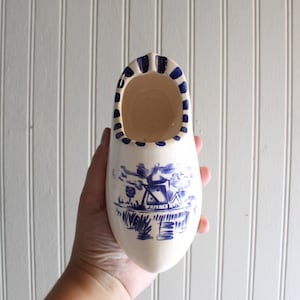 May include: A cream-colored ceramic clog featuring a hand-painted blue windmill landscape on the front and a series of blue vertical stripes decorating the inner rim. The shoe-shaped object has a smooth, glossy finish and a hollow interior.