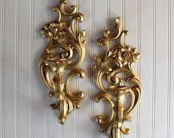May include: A pair of gold-colored wall sconces featuring intricate scrollwork and floral motifs. Each sconce has a central candle holder cup integrated into the ornate, elongated vertical frame with curved decorative elements and petals.