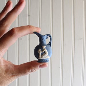 May include: A miniature blue ceramic pitcher featuring a white relief design of a kneeling figure. The small vessel has a curved handle, a flared spout, and decorative patterns around the neck and base, showcasing a classic matte finish.