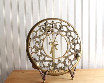 Puede incluir: Un salvamanteles circular de latón con un intrincado diseño calado de enredaderas y motivos florales. El centro muestra un ramo estilizado atado con un lazo. La pieza se apoya sobre un pequeño soporte de metal decorativo con pies texturizados.