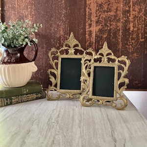 May include: Two ornate, gold-colored picture frames with black interiors sit on a light-colored surface. The frames have a detailed, scrollwork design. A brown and white pitcher with greenery and two green books are in the background.