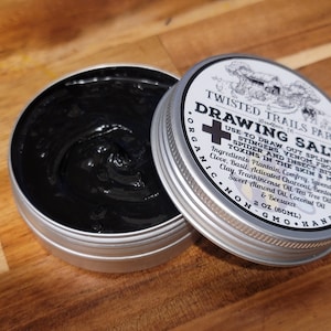 May include: A silver tin with a black label that reads "Twisted Trails Farm Drawing Salve" and lists ingredients and uses for the salve. The tin is open and a black salve is visible inside.