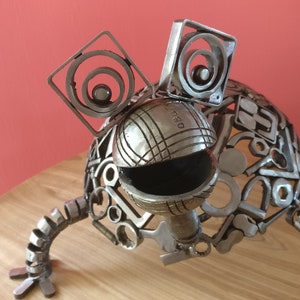 Peut inclure: Une sculpture en m&eacute;tal argent&eacute; d'une grenouille faite &agrave; partir de pi&egrave;ces de m&eacute;tal recycl&eacute;es. La grenouille a de grands yeux ronds et un corps rond textur&eacute;. La sculpture est pos&eacute;e sur une table en bois.