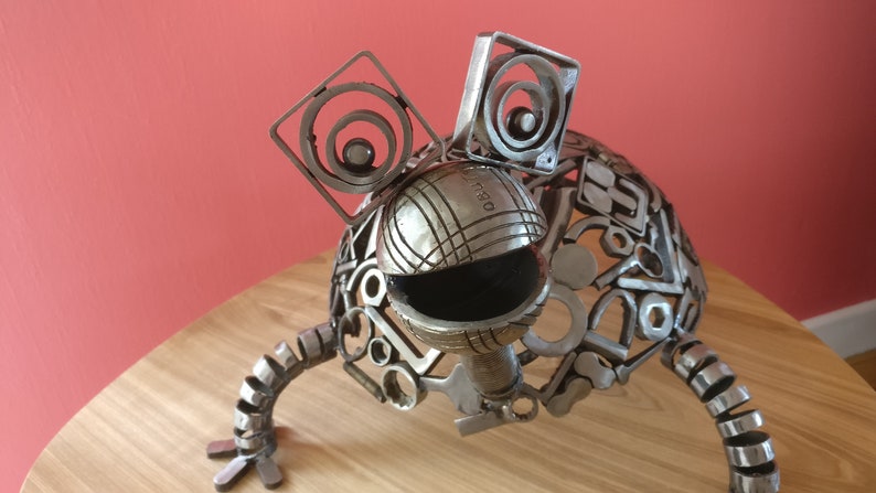 Peut inclure: Une sculpture en m&eacute;tal argent&eacute; d'une grenouille faite &agrave; partir de pi&egrave;ces de m&eacute;tal recycl&eacute;es. La grenouille a de grands yeux ronds et un corps rond textur&eacute;. La sculpture est pos&eacute;e sur une table en bois.