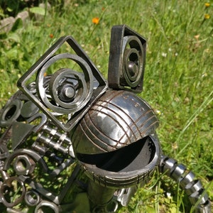 Peut inclure: Une sculpture de grenouille fantaisiste en m&eacute;tal, r&eacute;alis&eacute;e &agrave; partir de pi&egrave;ces de m&eacute;tal recycl&eacute;es. La grenouille a une grosse t&ecirc;te ronde avec une surface textur&eacute;e et deux grands yeux carr&eacute;s en cercles de m&eacute;tal. La grenouille est assise dans un lit d'herbe verte.