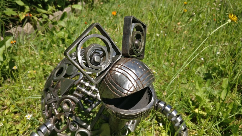 Peut inclure: Une sculpture de grenouille fantaisiste en m&eacute;tal, r&eacute;alis&eacute;e &agrave; partir de pi&egrave;ces de m&eacute;tal recycl&eacute;es. La grenouille a une grosse t&ecirc;te ronde avec une surface textur&eacute;e et deux grands yeux carr&eacute;s en cercles de m&eacute;tal. La grenouille est assise dans un lit d'herbe verte.