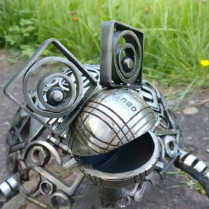 Peut inclure: Une sculpture de grenouille en m&eacute;tal faite &agrave; partir de mat&eacute;riaux recycl&eacute;s, y compris une boule de p&eacute;tanque. La grenouille a une grosse t&ecirc;te ronde avec une bouche noire et deux oreilles faites de bagues m&eacute;talliques. Le corps de la grenouille est fait de diverses pi&egrave;ces de m&eacute;tal, y compris des &eacute;crous, des boulons et des engrenages.