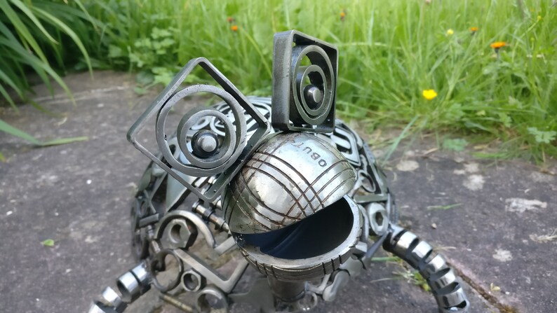 Peut inclure: Une sculpture de grenouille en m&eacute;tal faite &agrave; partir de mat&eacute;riaux recycl&eacute;s, y compris une boule de p&eacute;tanque. La grenouille a une grosse t&ecirc;te ronde avec une bouche noire et deux oreilles faites de bagues m&eacute;talliques. Le corps de la grenouille est fait de diverses pi&egrave;ces de m&eacute;tal, y compris des &eacute;crous, des boulons et des engrenages.