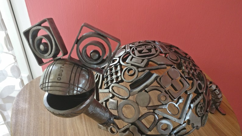 Peut inclure: Une sculpture en m&eacute;tal d'une tortue r&eacute;alis&eacute;e &agrave; partir de diverses pi&egrave;ces de m&eacute;tal recycl&eacute;es. La tortue a une carapace ronde et une t&ecirc;te avec deux grands yeux. La sculpture est peinte en argent et porte l'inscription "OBUT" sur la t&ecirc;te.