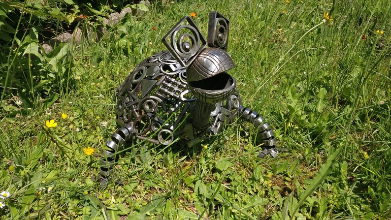 Peut inclure: Une sculpture de grenouille en m&eacute;tal faite &agrave; partir de pi&egrave;ces de m&eacute;tal recycl&eacute;es. La grenouille est assise sur un lit d'herbe verte.