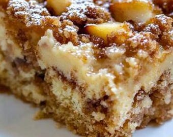 May include: A close-up of a slice of apple crumble cake on a white plate. The cake has a golden-brown crumb topping, chunks of apple, and a creamy layer. Powdered sugar dusts the top, enhancing the dessert's appeal.