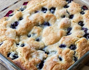 May include: A baked blueberry cobbler in a clear glass baking dish. The cobbler has a golden-brown, slightly textured crust with visible blueberries throughout. The dish sits on a light-colored wooden surface.