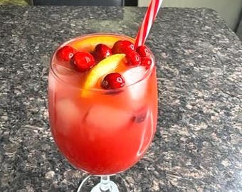 May include: A vibrant red cocktail in a stemmed glass, garnished with cranberries and a lemon slice. A red and white striped straw extends from the drink. The glass sits on a speckled countertop.