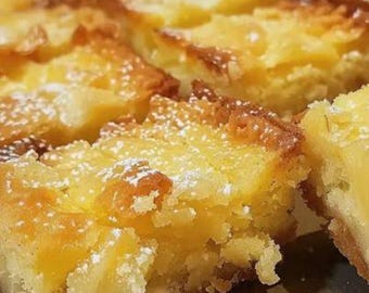 May include: Close-up of several square-cut lemon bars dusted with powdered sugar. The bars have a golden-brown, slightly crispy top layer and a bright yellow, moist filling. The dessert is arranged on a reflective surface.