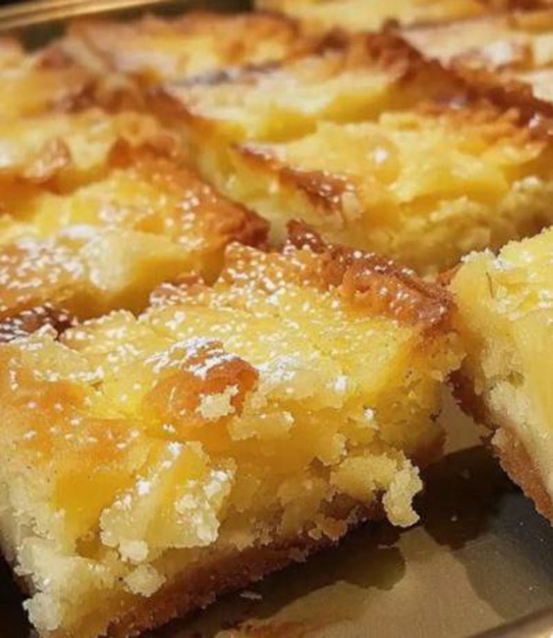 May include: Close-up of several square-cut lemon bars dusted with powdered sugar. The bars have a golden-brown, slightly crispy top layer and a bright yellow, moist filling. The dessert is arranged on a reflective surface.