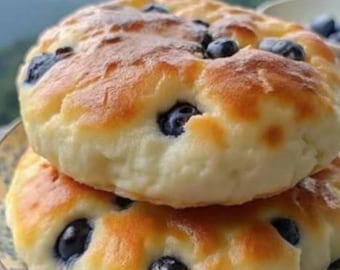 May include: Two round, golden-brown baked bread rolls stacked on top of each other. The surface of the bread is soft and pale yellow, featuring several dark blue, whole blueberries embedded into the dough. The crust shows light brown toasted spots.