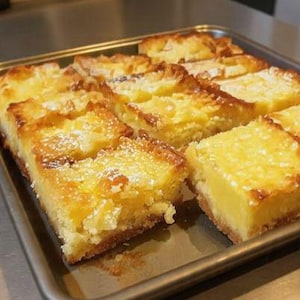 Peut inclure: Une plaque de barres au citron fraîchement cuites. Les barres sont jaune doré avec une croûte friable et saupoudrées de sucre glace. Le plateau est de couleur métal foncé.