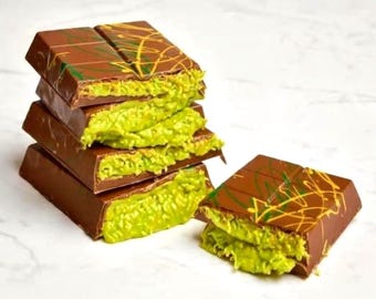 May include: A stack of four rectangular milk chocolate bars and one separate bar, each featuring a smooth brown exterior with thin yellow and green decorative drizzles. The bars are broken open to reveal a textured, bright lime green filling.
