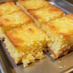 May include: A tray of golden-brown, square-cut baked goods. The tops have a glossy, caramelized appearance, and the edges are slightly darker. The texture appears moist and crumbly, suggesting a dessert like a bar or cake.